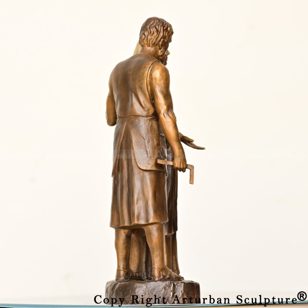 Back-angle view of a bronze Mary and Joseph statue, showing Joseph holding a carpenter's square in his right hand behind Mary